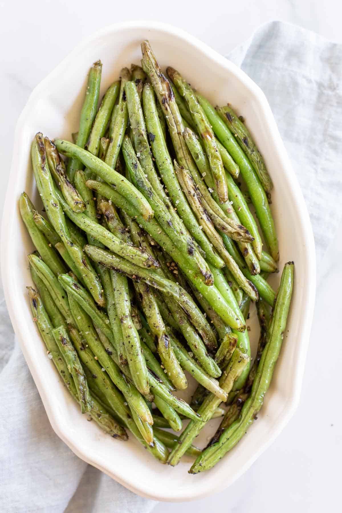 A white oval serving dish, filled with grilled green beans, blue linen towel to the side.