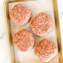 A gold baking sheet with four burger patties on parchment paper.