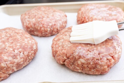 A gold baking sheet with four burger patties on parchment paper, a brush brushing olive oil on one.