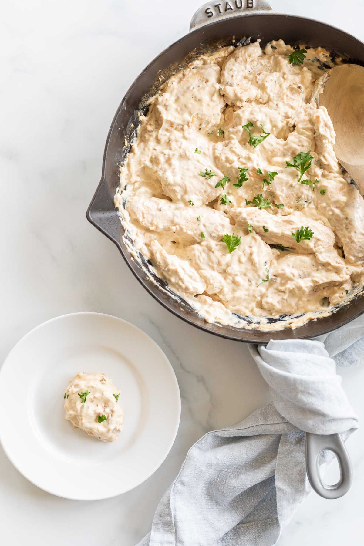 A skillet filled with cream cheese chicken, with a small serving on a white plate in foreground