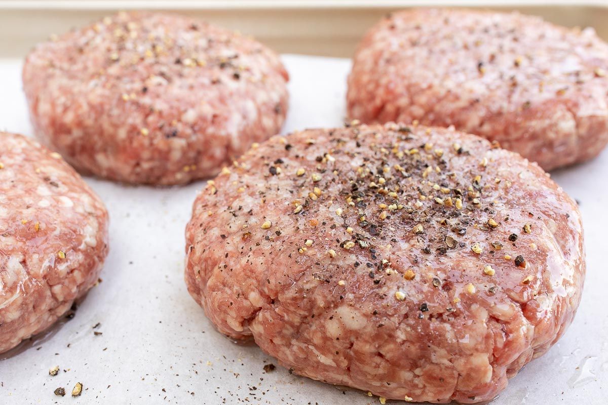 Homemade burger patties, uncooked, topped with seasoning on parchment paper.