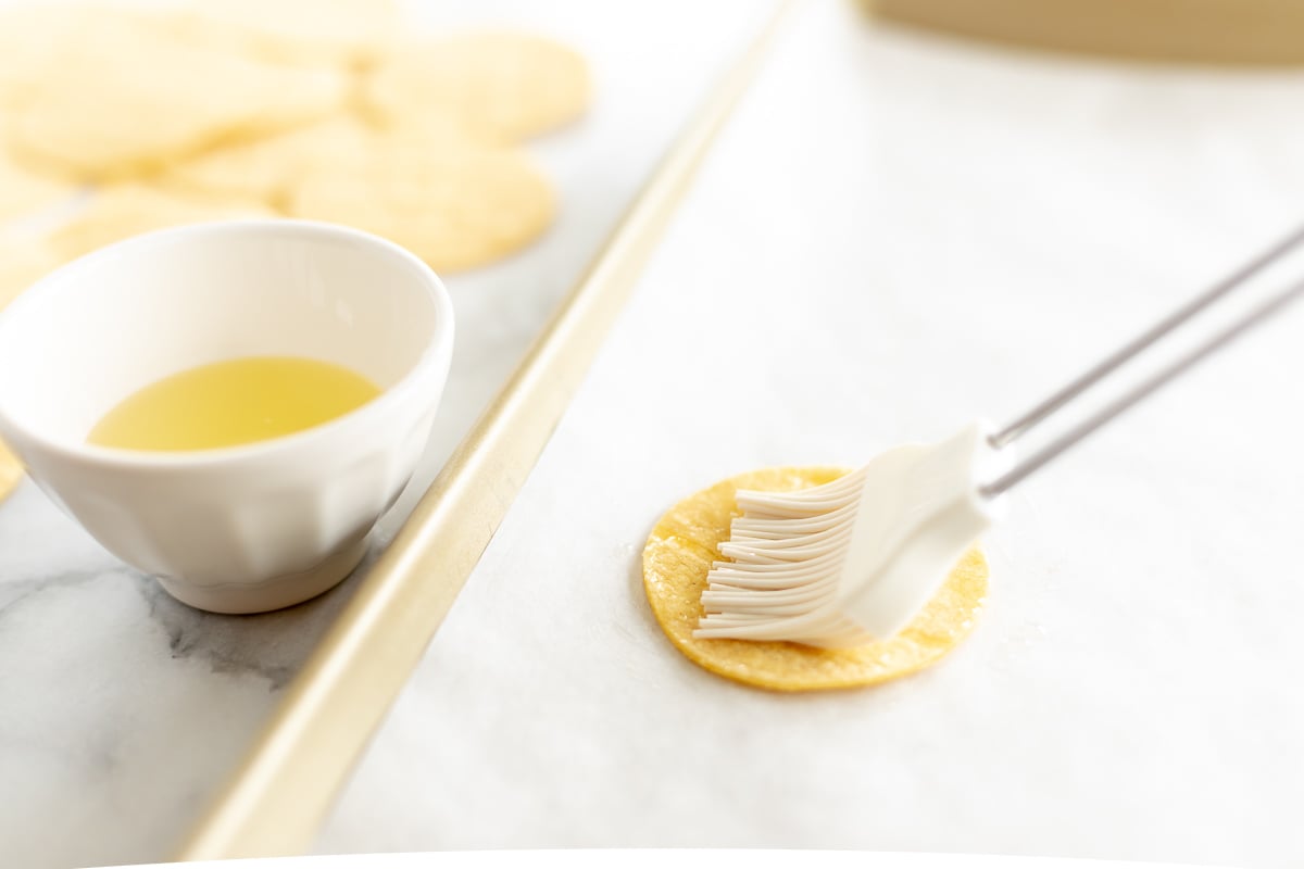 A corn tortilla being brushed with oil on a baking sheet.