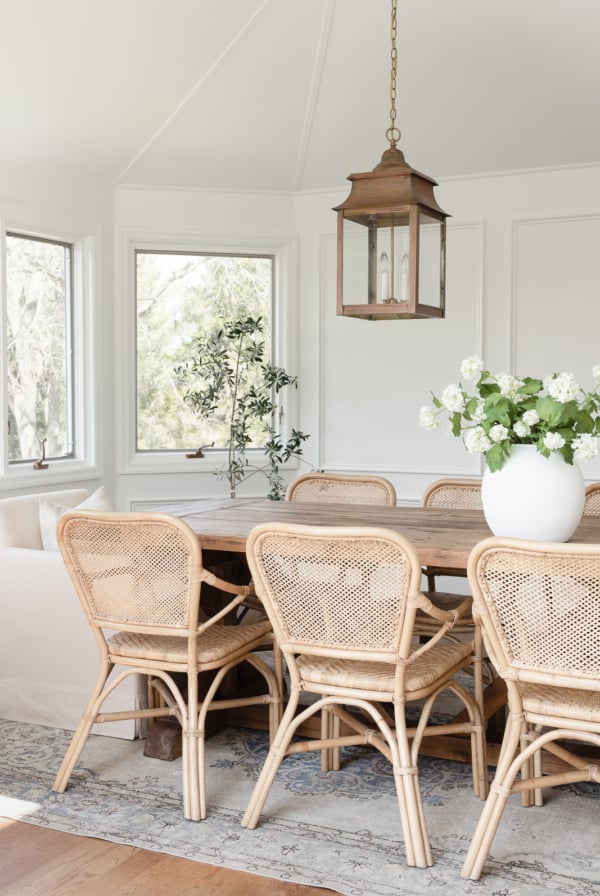 A white dining room with a wood table, rattan chairs and picture frame moulding on the walls.