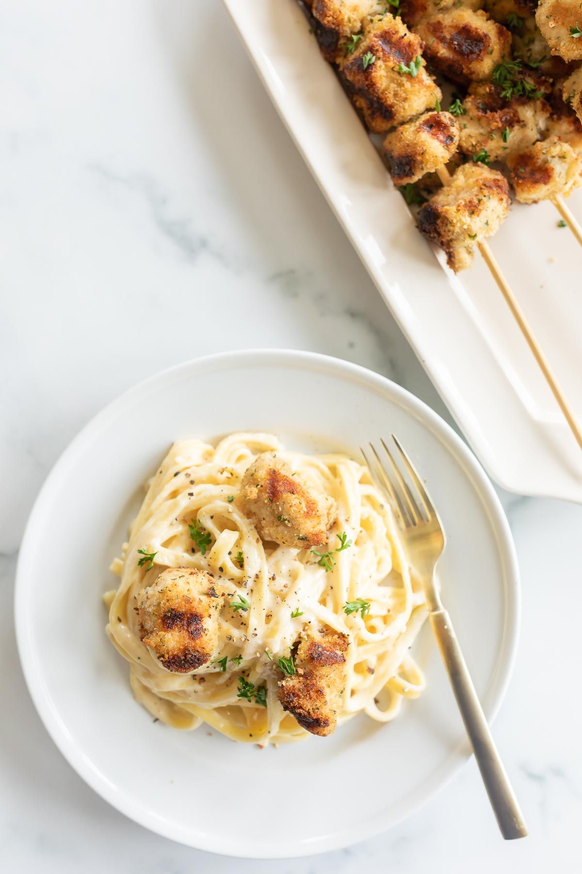 A white plate with fettuccine alfredo topped with spiedini, platter of chicken spiedini in the background.