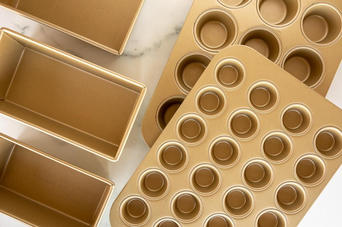 A variety of gold cake pans, loaf pans and muffin baking pan sizes laid out on a marble surface.