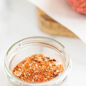 A small glass bowl of burger seasoning, with an uncooked burger patty to the side