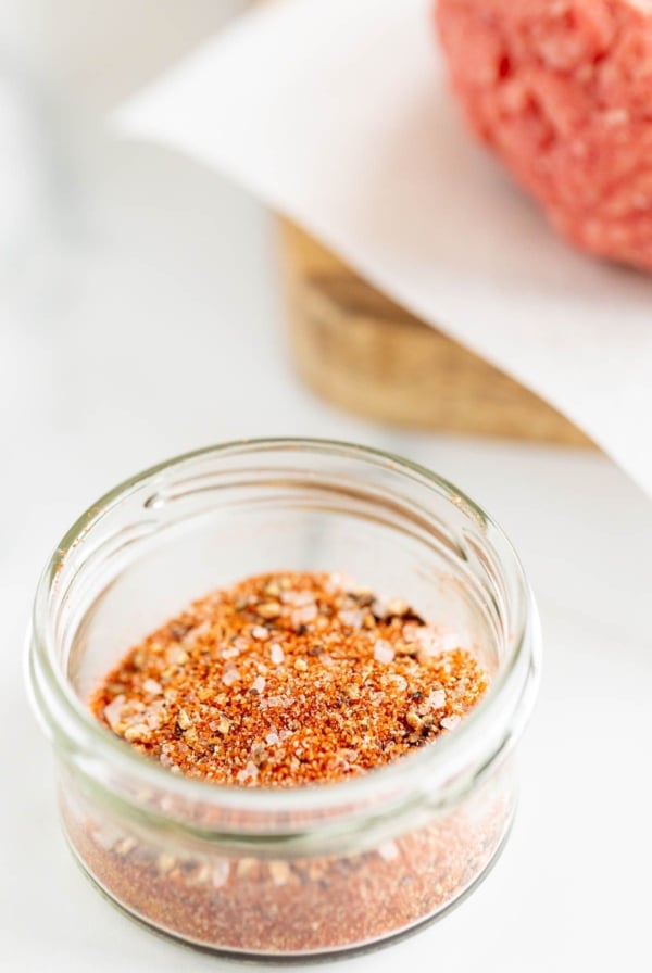 A small glass bowl of burger seasoning, with an uncooked burger patty to the side