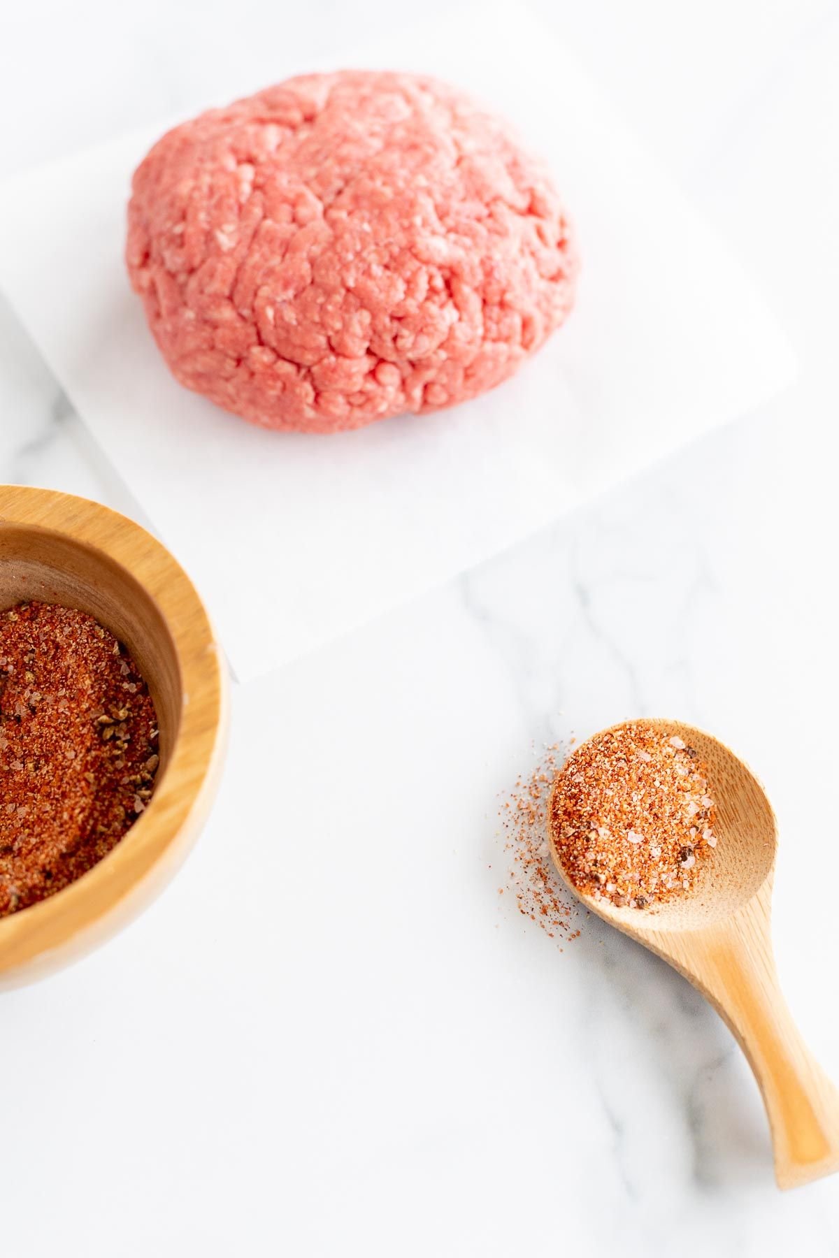 A small wooden bowl of burger seasoning, with an uncooked burger patty to the side