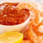 An oval platter of roasted shrimp cocktail with a homemade cocktail sauce in a white bowl.