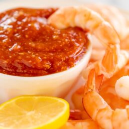 An oval platter of roasted shrimp cocktail with a homemade cocktail sauce in a white bowl.