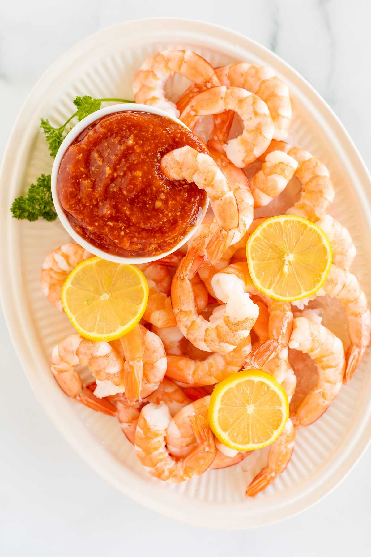 An oval platter of roasted shrimp cocktail with a homemade cocktail sauce in a white bowl.