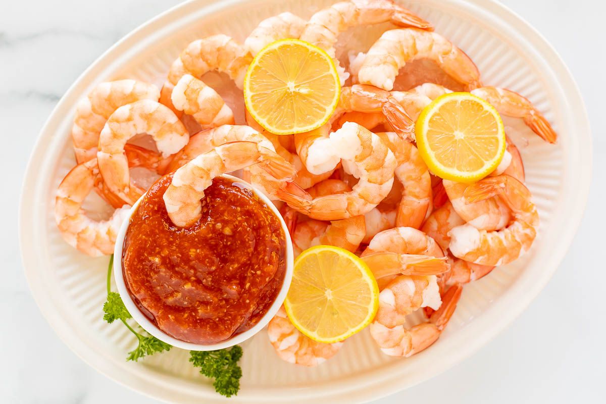 An oval platter of roasted shrimp cocktail with a homemade cocktail sauce in a white bowl.