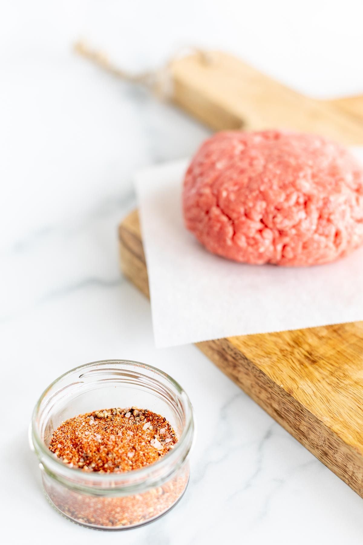 A small glass bowl of burger seasoning, with an uncooked burger patty to the side