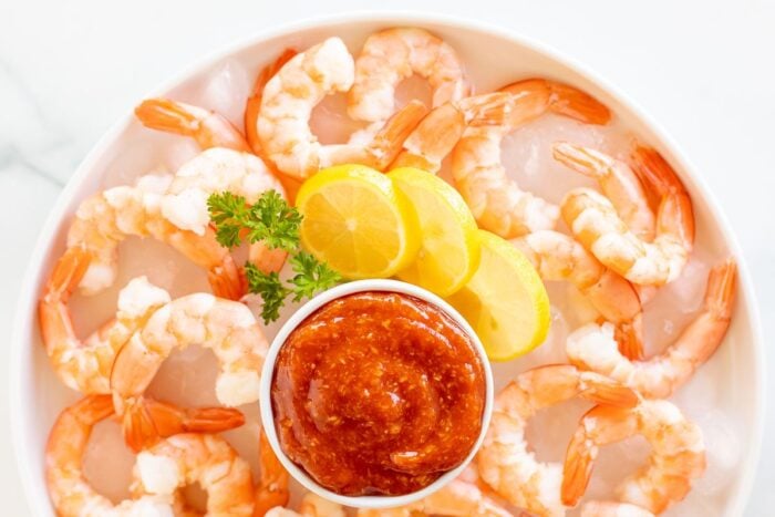 An oval platter of roasted shrimp cocktail with a homemade cocktail sauce in a white bowl.