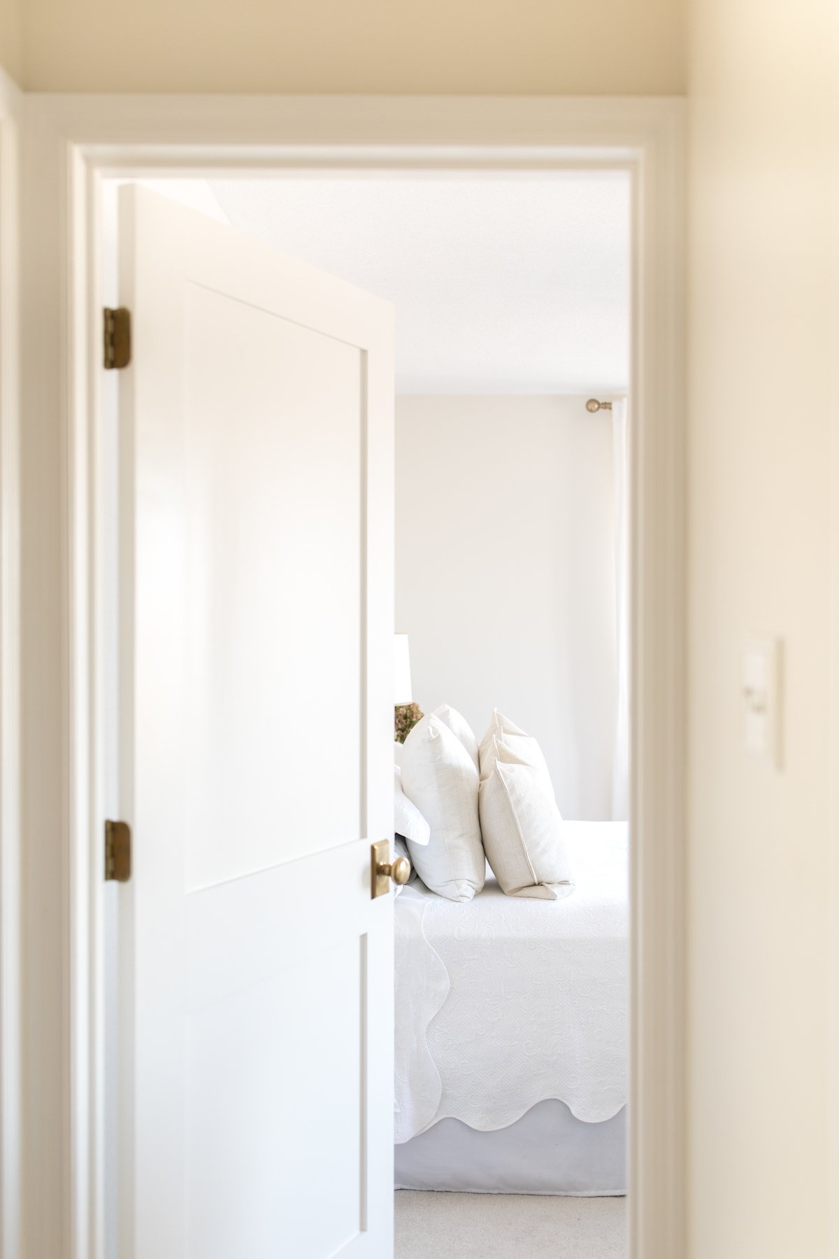 A bright and airy bedroom viewed through an open door, featuring a painting hanging over the interior side of the door.