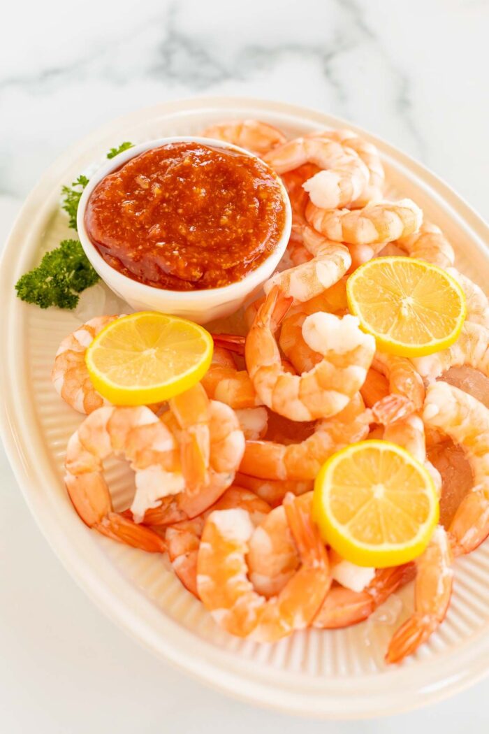 An oval platter of roasted shrimp cocktail with a homemade cocktail sauce in a white bowl.