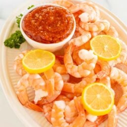 An oval platter of roasted shrimp cocktail with a homemade cocktail sauce in a white bowl.