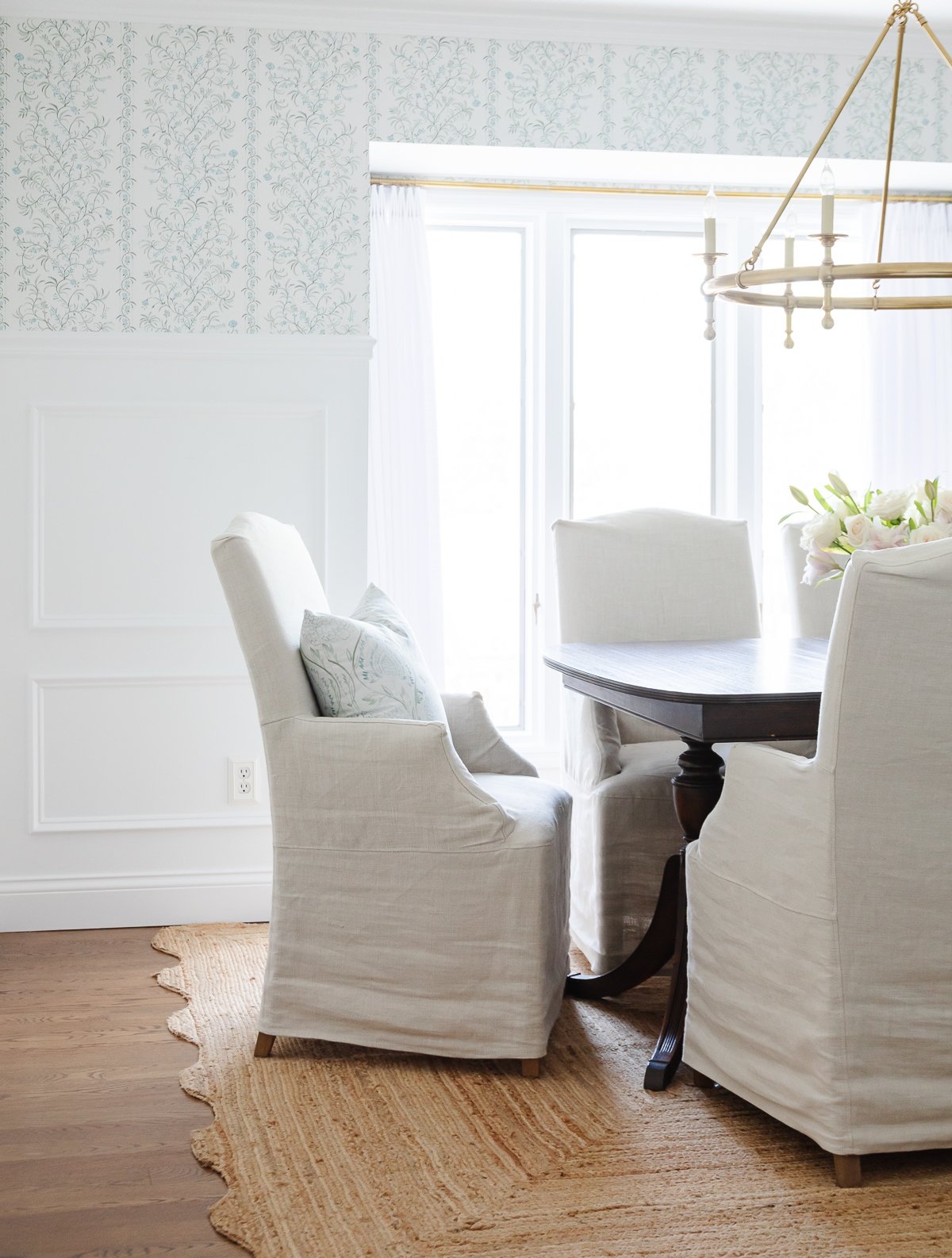 Elegant dining room featuring a dark wood table, white slipcovered chairs, a beige rug, and a gold chandelier, with Benjamin Moore Chantilly Lace walls and floral wallpaper.