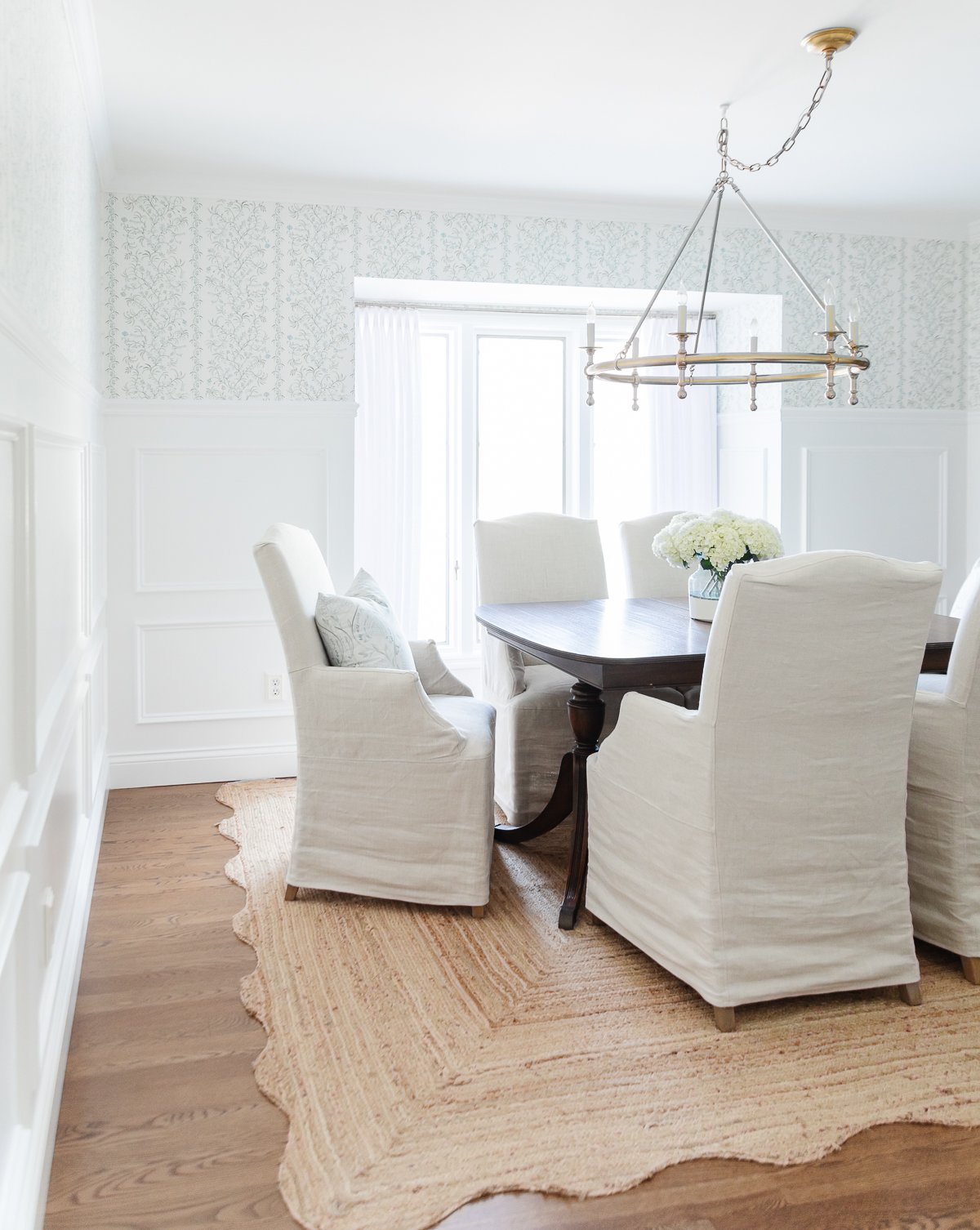 Elegant dining room featuring a dark wood table, white slipcovered chairs, a beige rug, and a gold chandelier, with Benjamin Moore Chantilly Lace walls and floral wallpaper. 