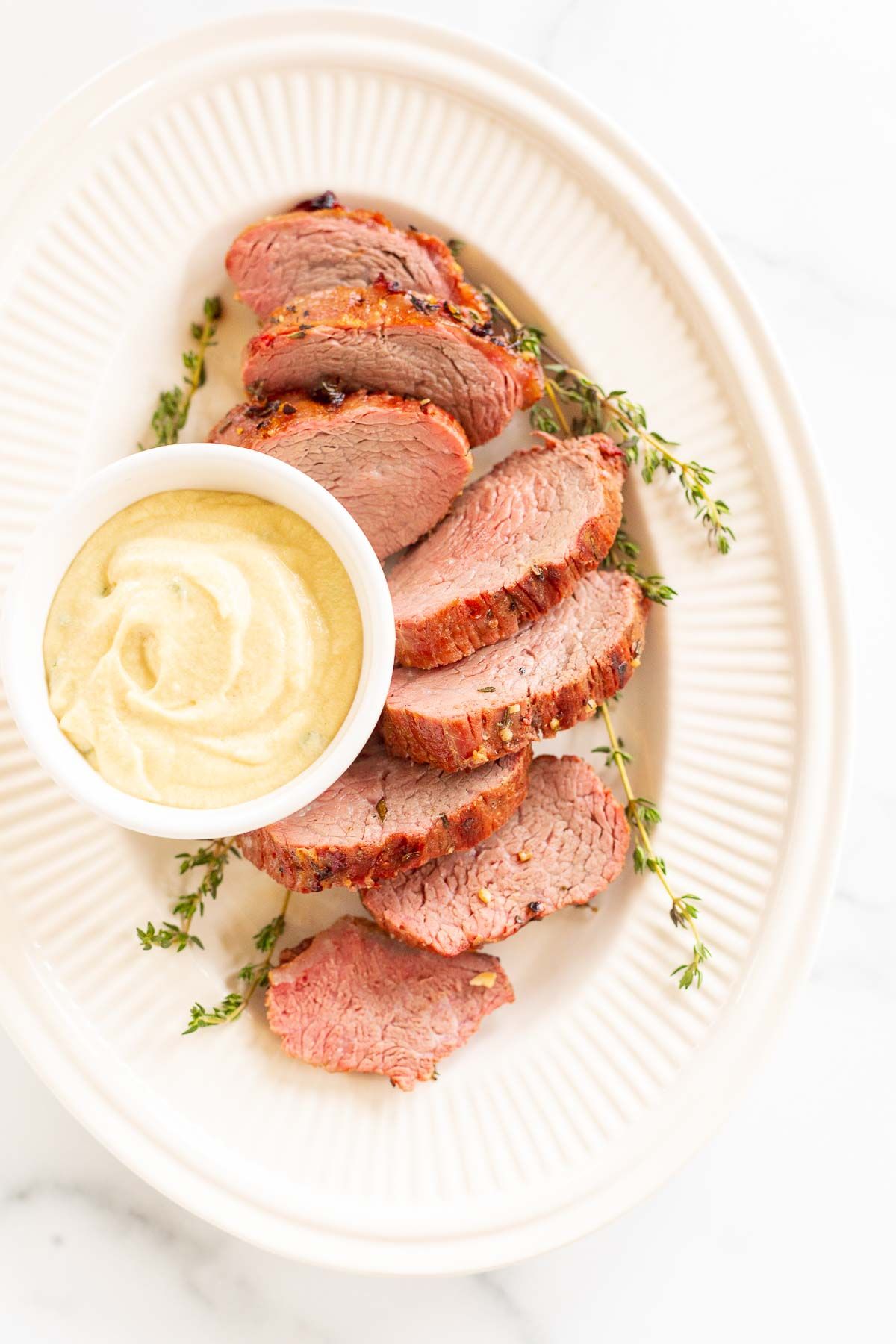 A white bowl full of Dijon mustard sauce, surrounded by beef tenderloin on a platter