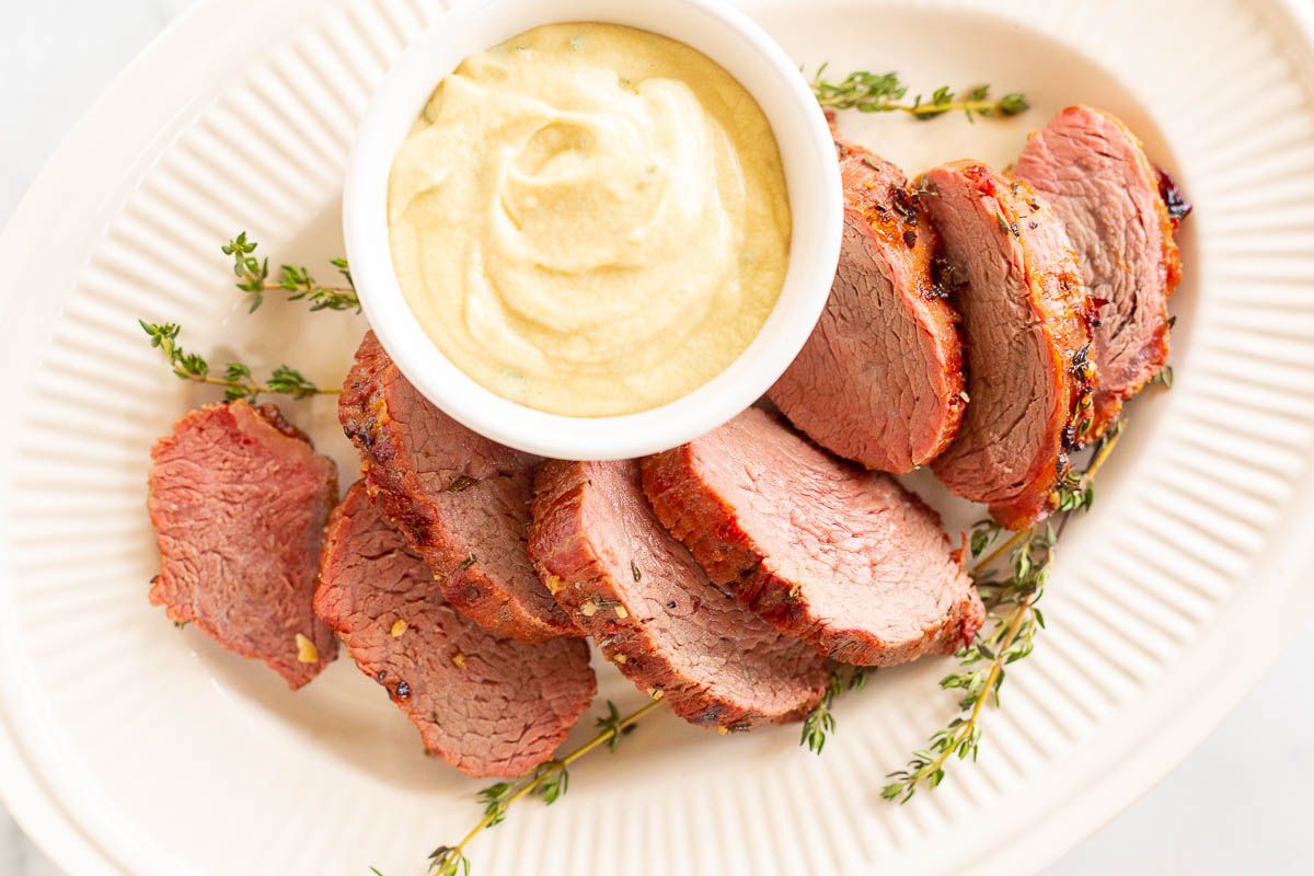 A white bowl full of Dijon mustard sauce, surrounded by beef tenderloin on a platter