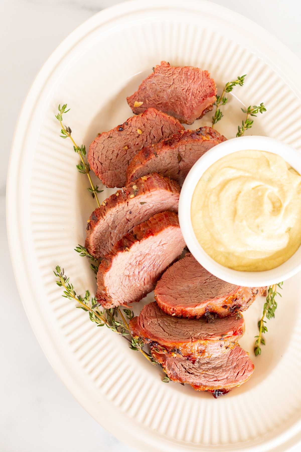 A white bowl full of Dijon mustard sauce, surrounded by beef tenderloin on a platter