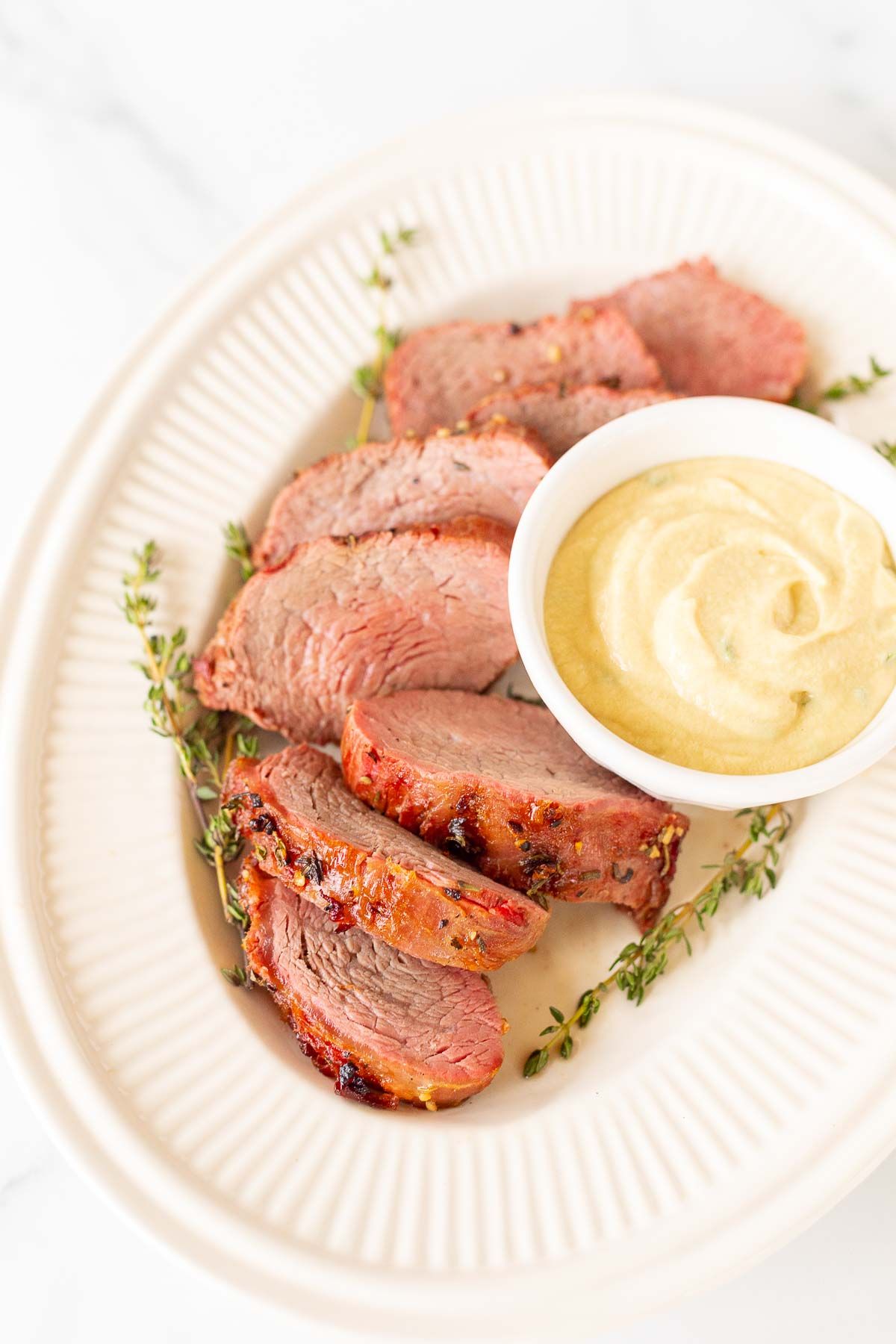 A white bowl full of Dijon mustard sauce, surrounded by beef tenderloin on a platter