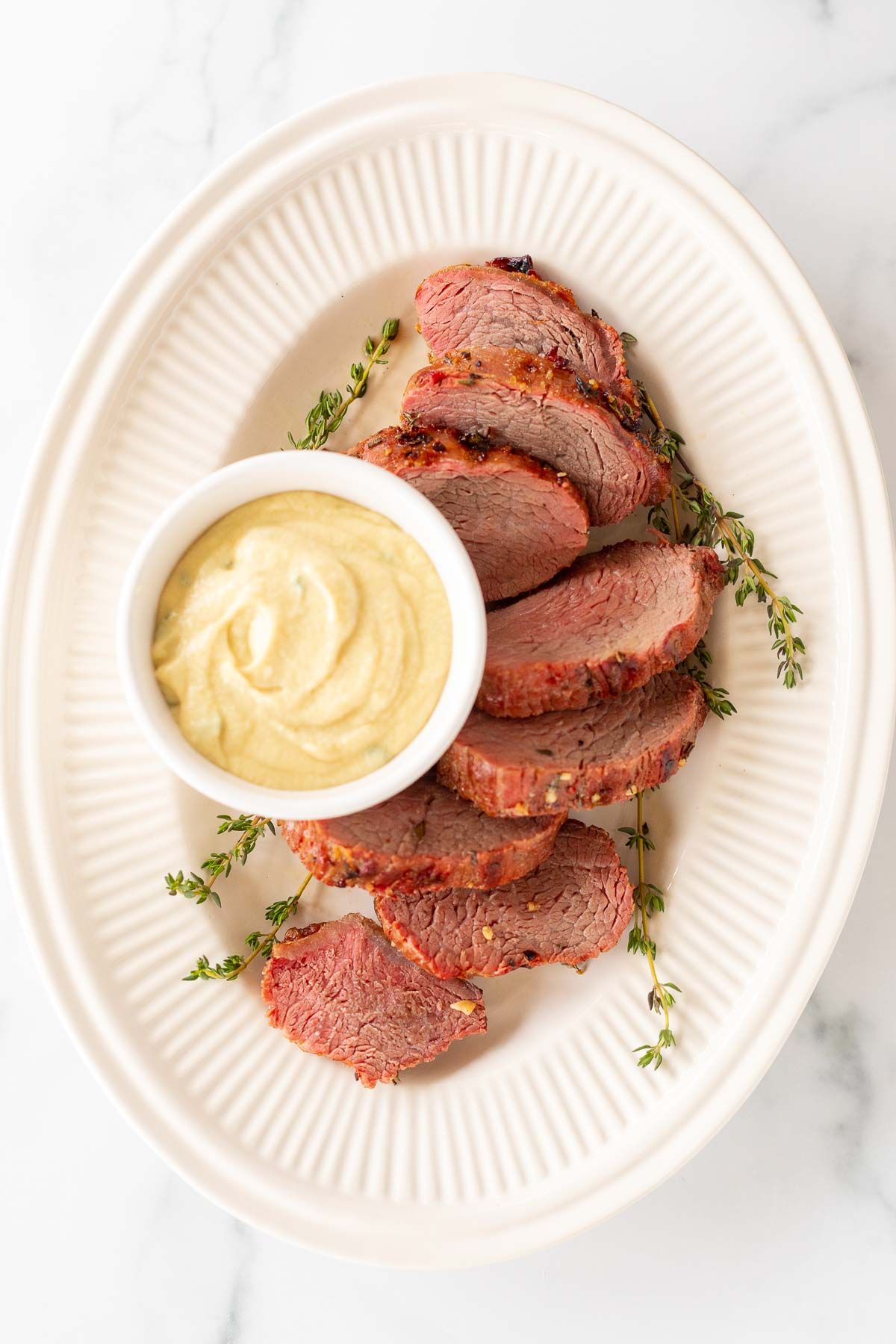 A white bowl full of Dijon mustard sauce, surrounded by beef tenderloin on a platter