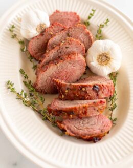 A platter of sliced beef tenderloin, garnished with fresh thyme and garlic.