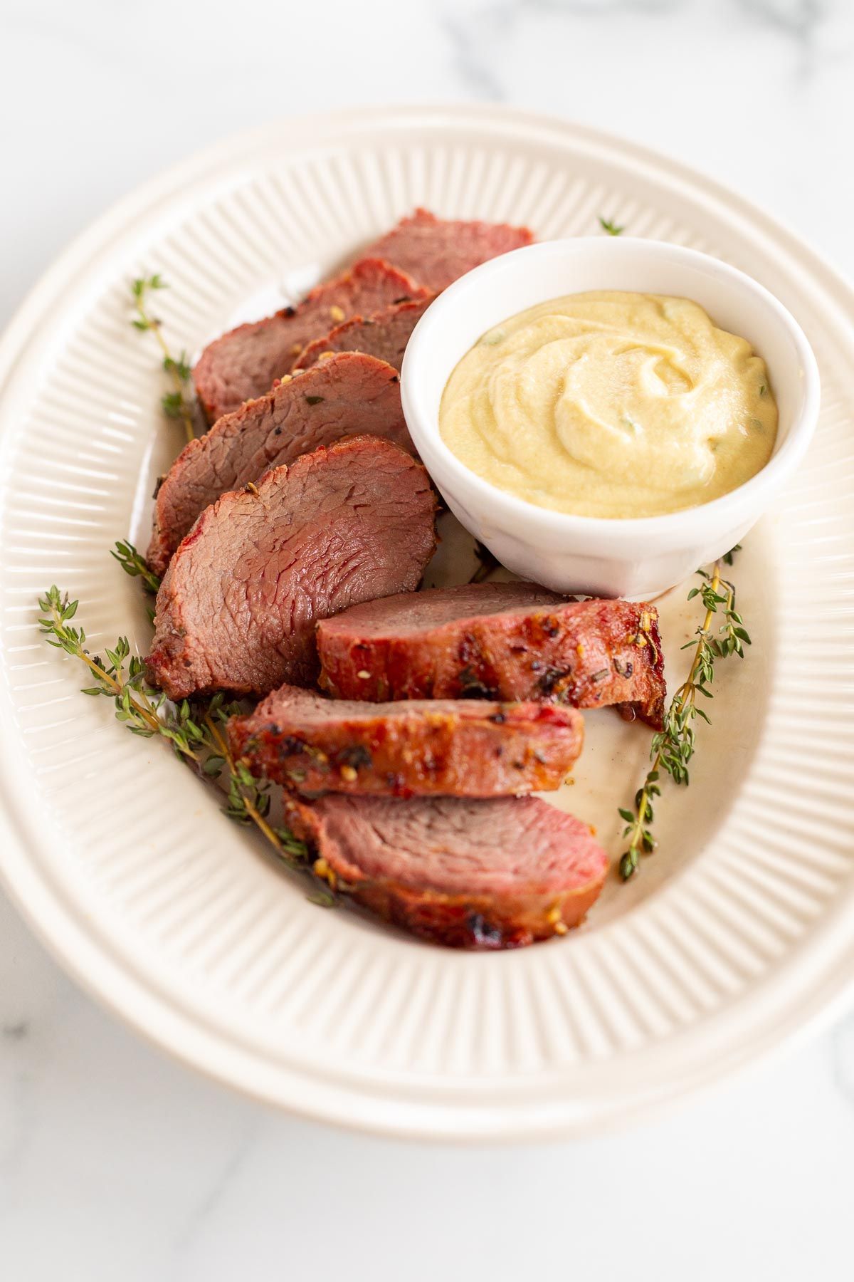 A grilled beef tenderloin on an oval platter with a bowl of dijon mustard sauce.