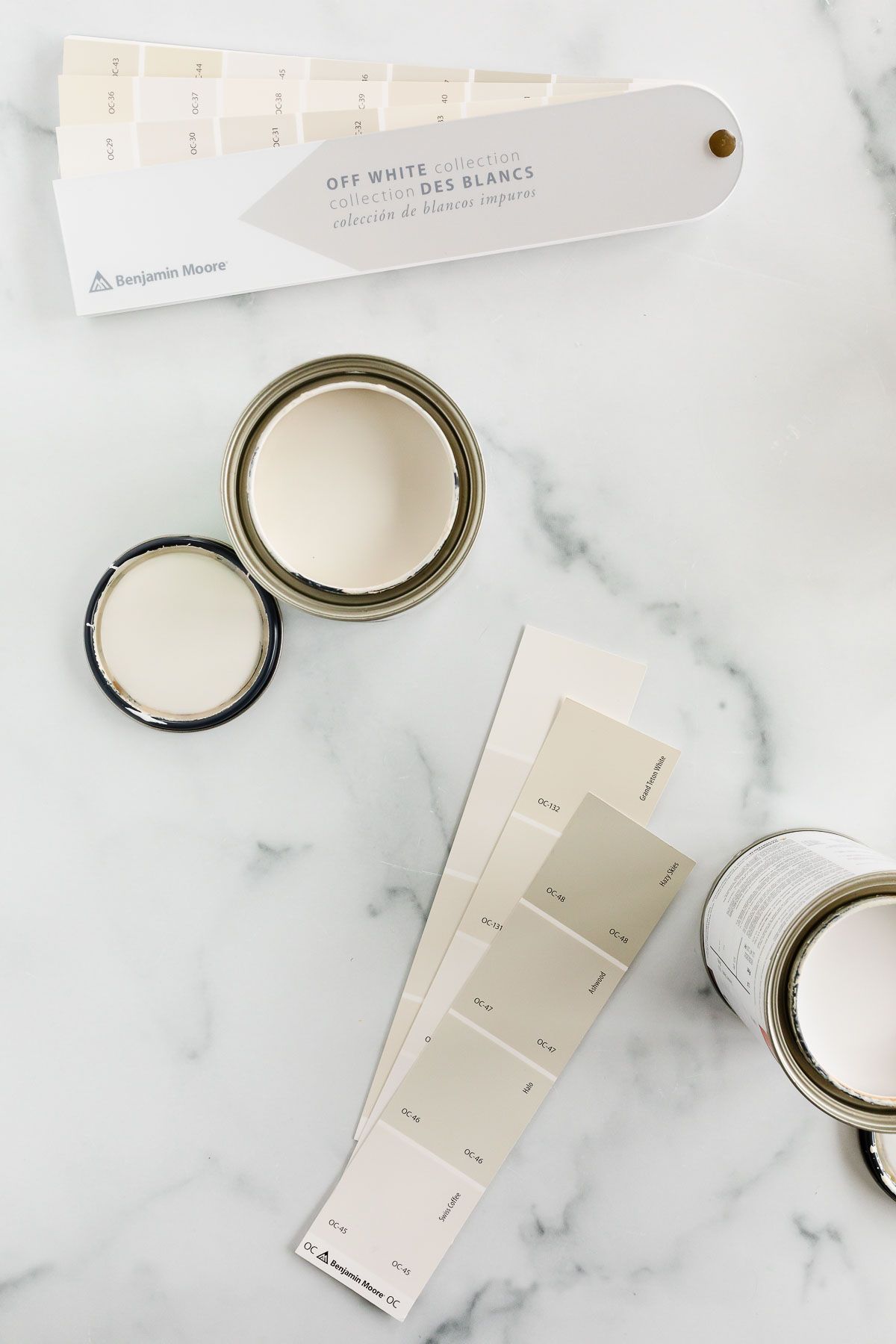 Open cans of warm whites and paint swatch cards laid out on a marble surface.