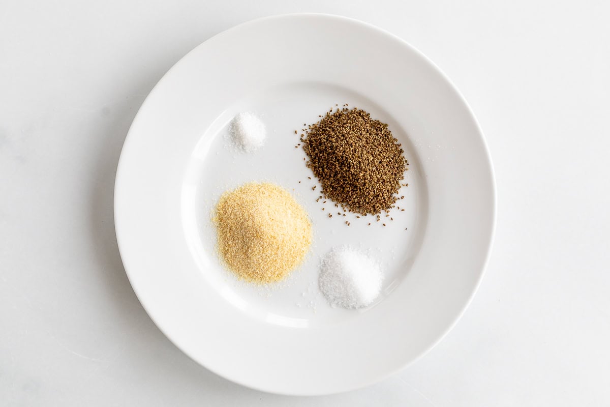 A white plate holds a simple composition of spices: salt, brown seeds, granulated yellow, and a fine white powder, reminiscent of beau monde seasoning's balanced elegance.