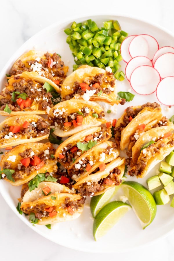 Plate of tacos with ground beef, cheese, tomatoes, and cilantro, served with sliced radishes, cucumbers, and lime wedges on a white surface for a Cinco de Mayo menu