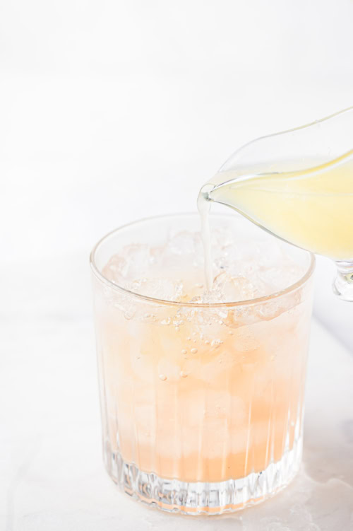 Lime juice being poured into a paloma cocktail glass filled with ice cubes on a white background.