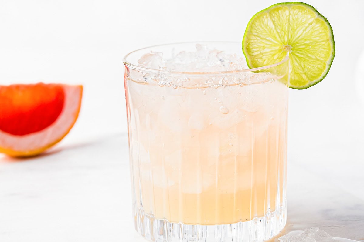 A glass of pink paloma soda with ice, garnished with a lime slice, on a white background with a grapefruit slice visible.