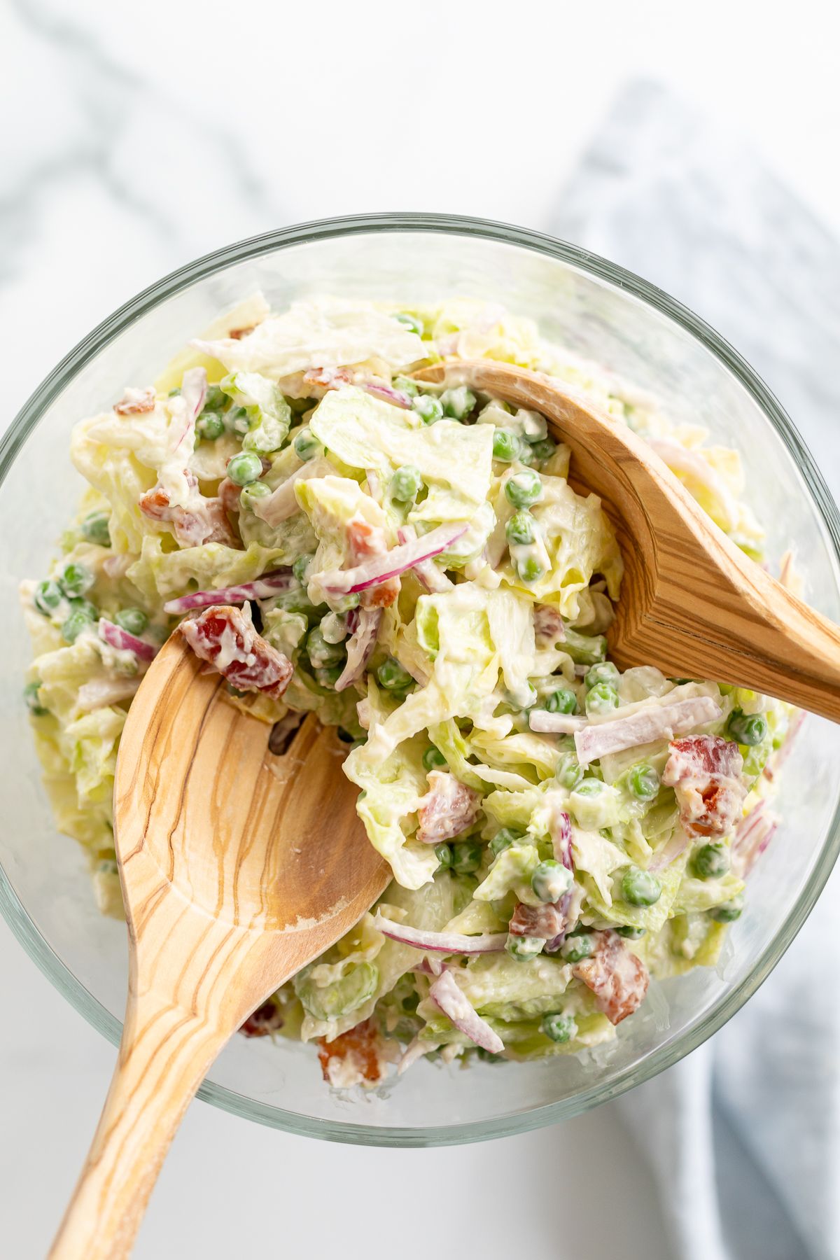 A 7 layer salad mixed up in a glass serving dish, wooden spoons to the side