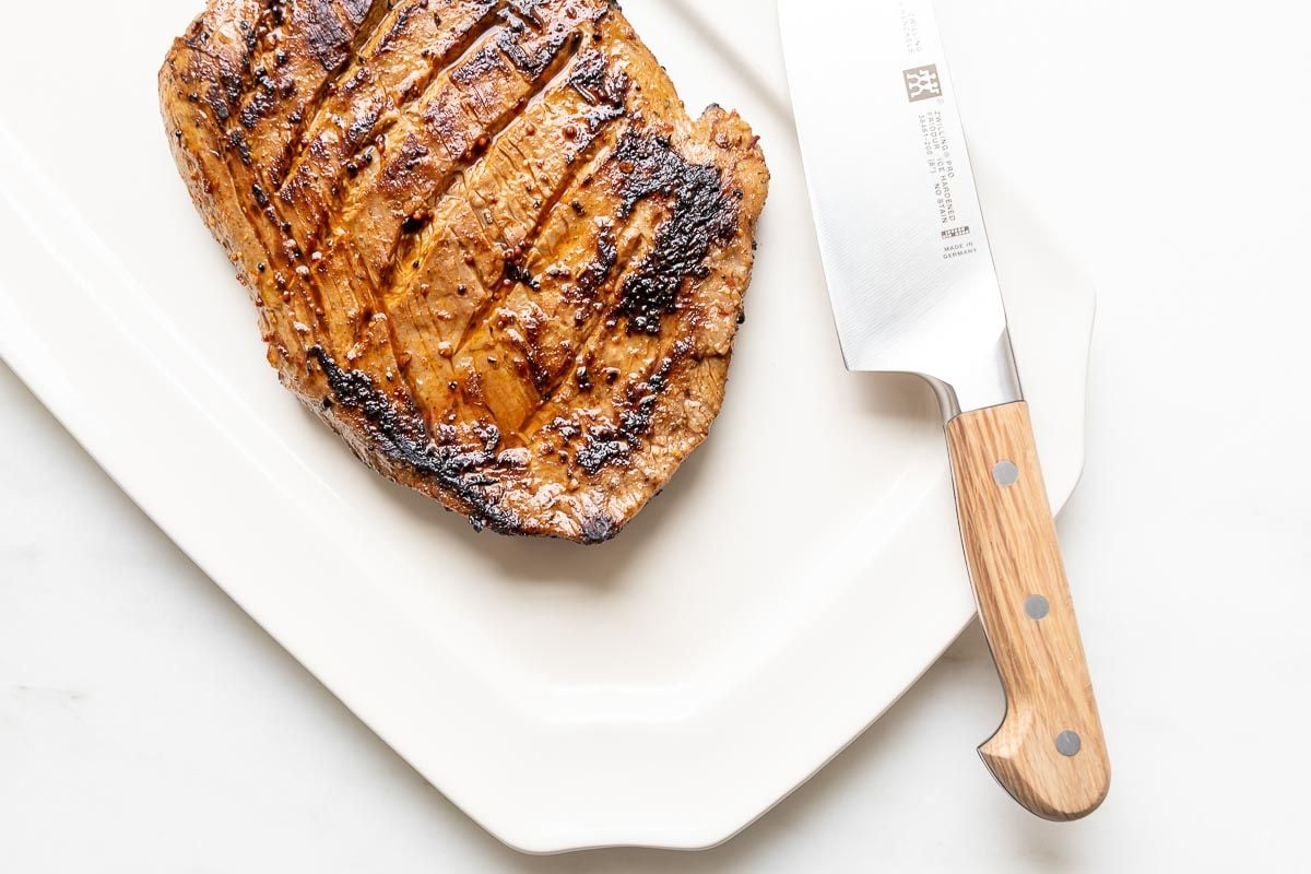 A chunk of cooked carne asada on a white platter,knife to the side