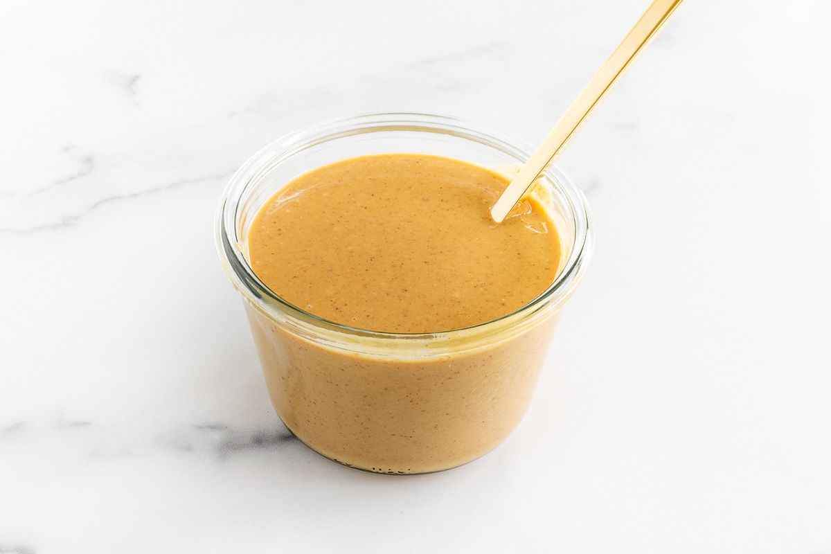 A small glass jar of homemade peanut butter on a marble surface, gold spoon to the side.