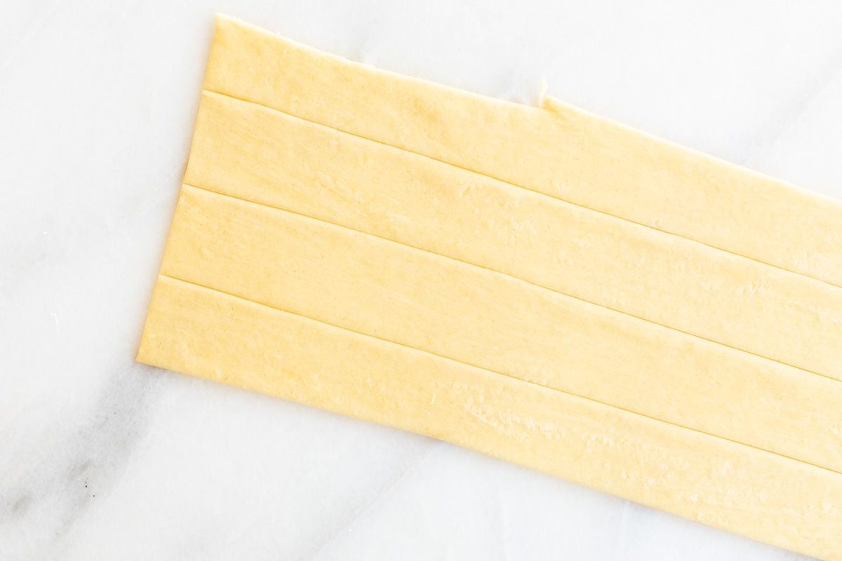 A sheet of homemade pasta dough laid out on a marble surface, cut into strips for Pappardelle