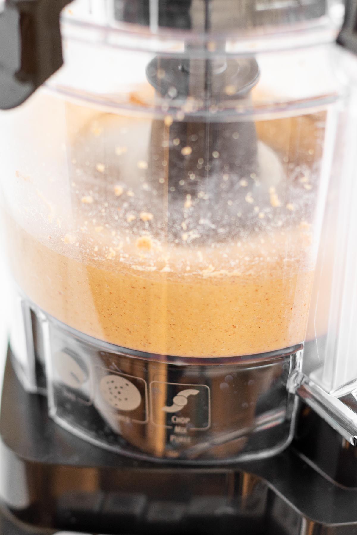 Image features homemade peanut butter in a food processor as part of a tutorial for how to make peanut butter.