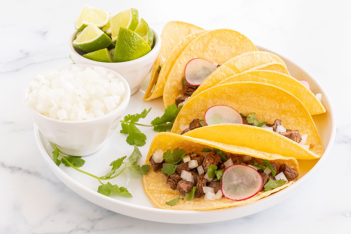 Street tacos on a white platter with a bowl of sliced limes and sliced onions