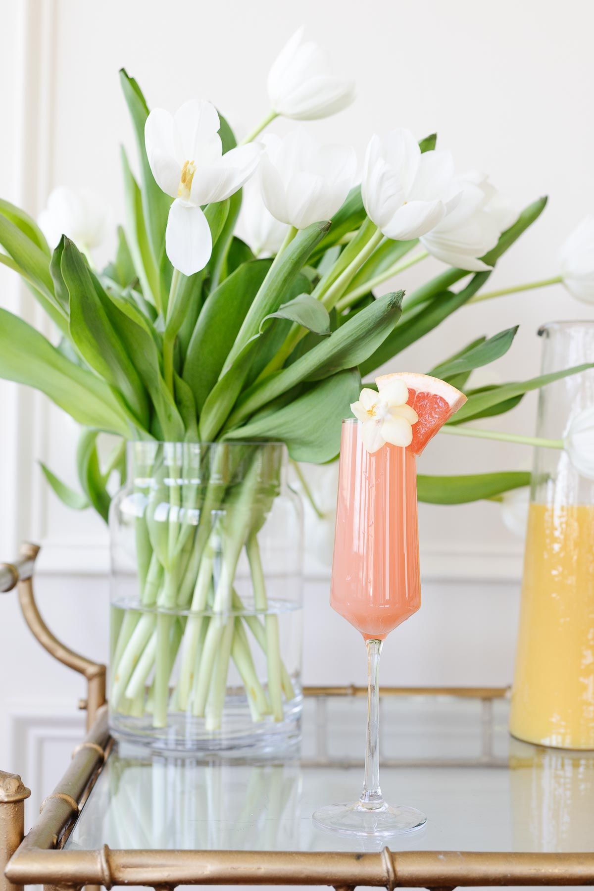 A grapefruit paloma on a gold bar cart, white tulips and fresh juices in the background