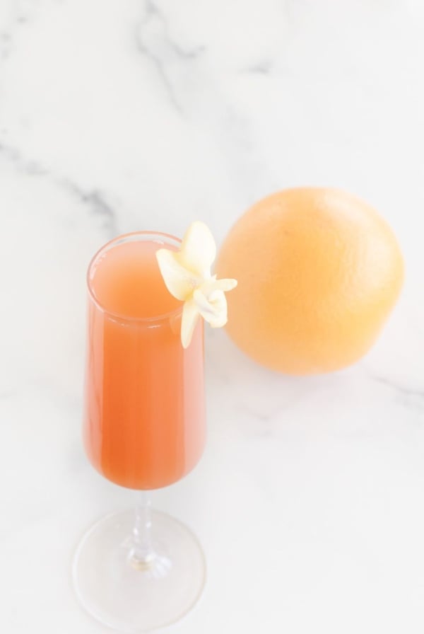 A grapefruit mimosa on a marble surface, garnished with a flower and a whole grapefruit in the background