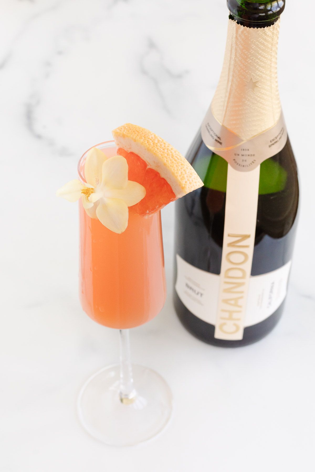 A grapefruit paloma on a marble surface, bottle of champagne to the side