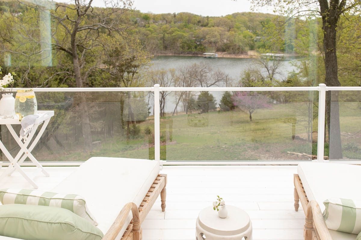 A white deck with glass railing, with two wooden lounge chairs and a ceramic garden stool in between.