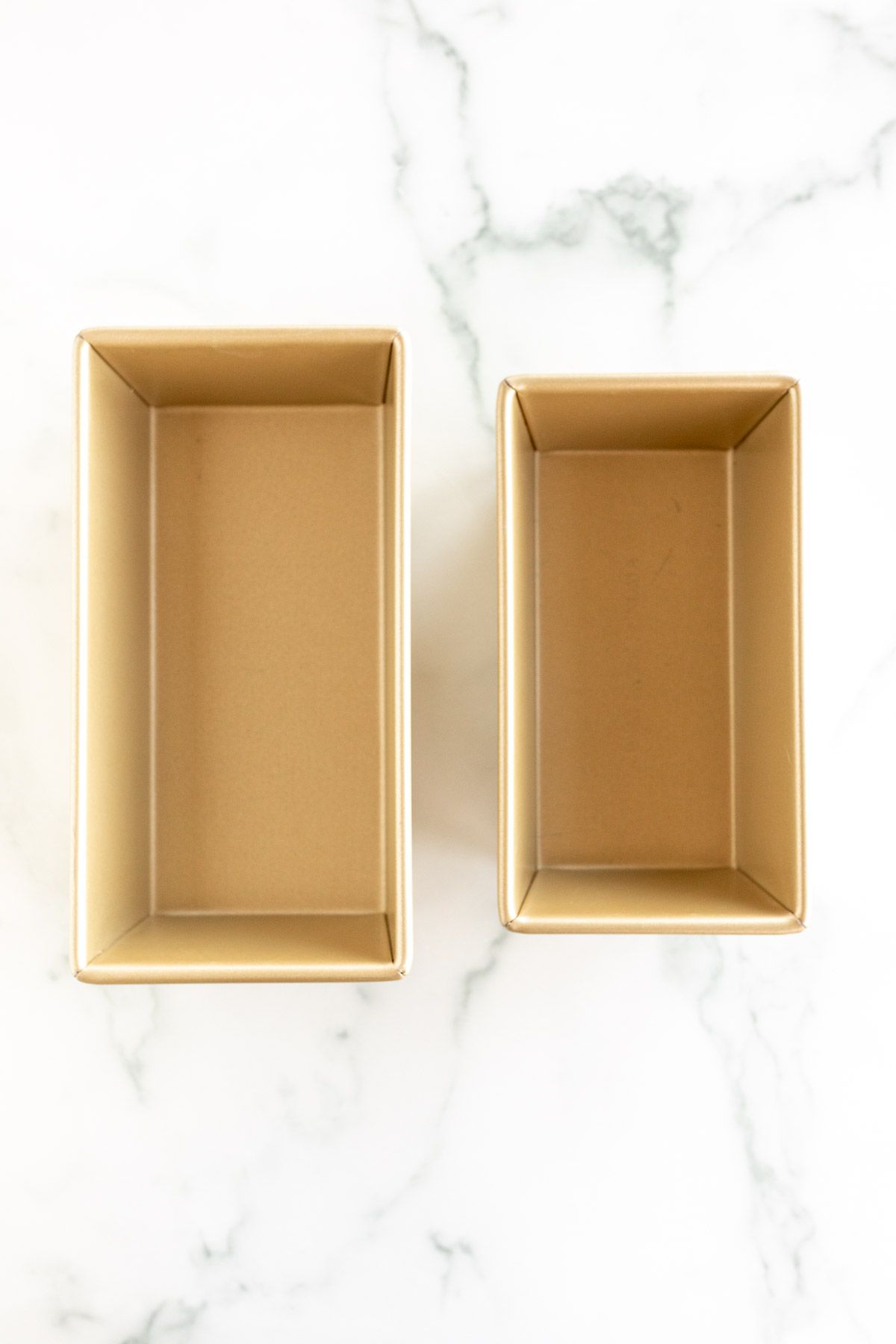 Two different gold loaf pan sizes on a white marble surface