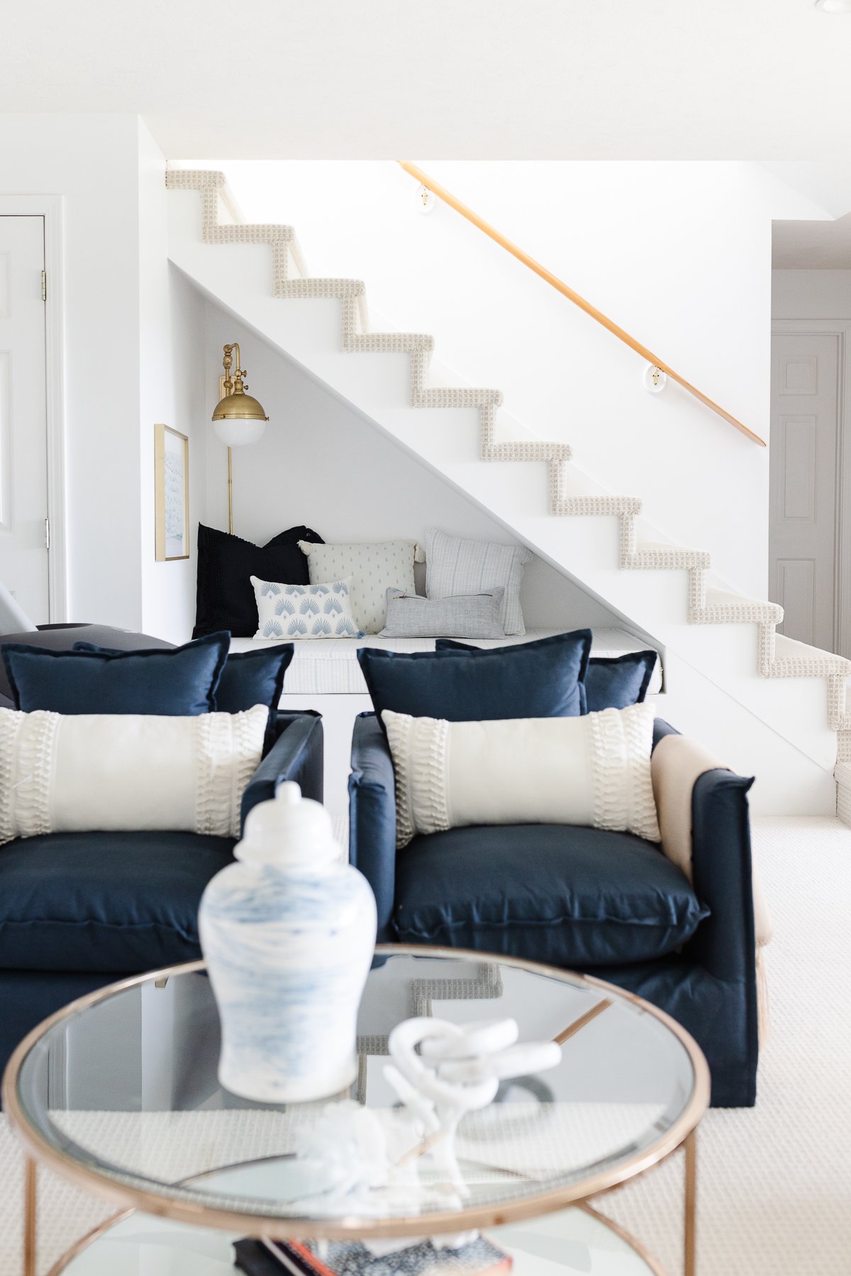 A well-illuminated living room with a white color scheme, featuring dark blue sofas, a glass coffee table of the standard coffee table height, and stairs leading upwards to the left.