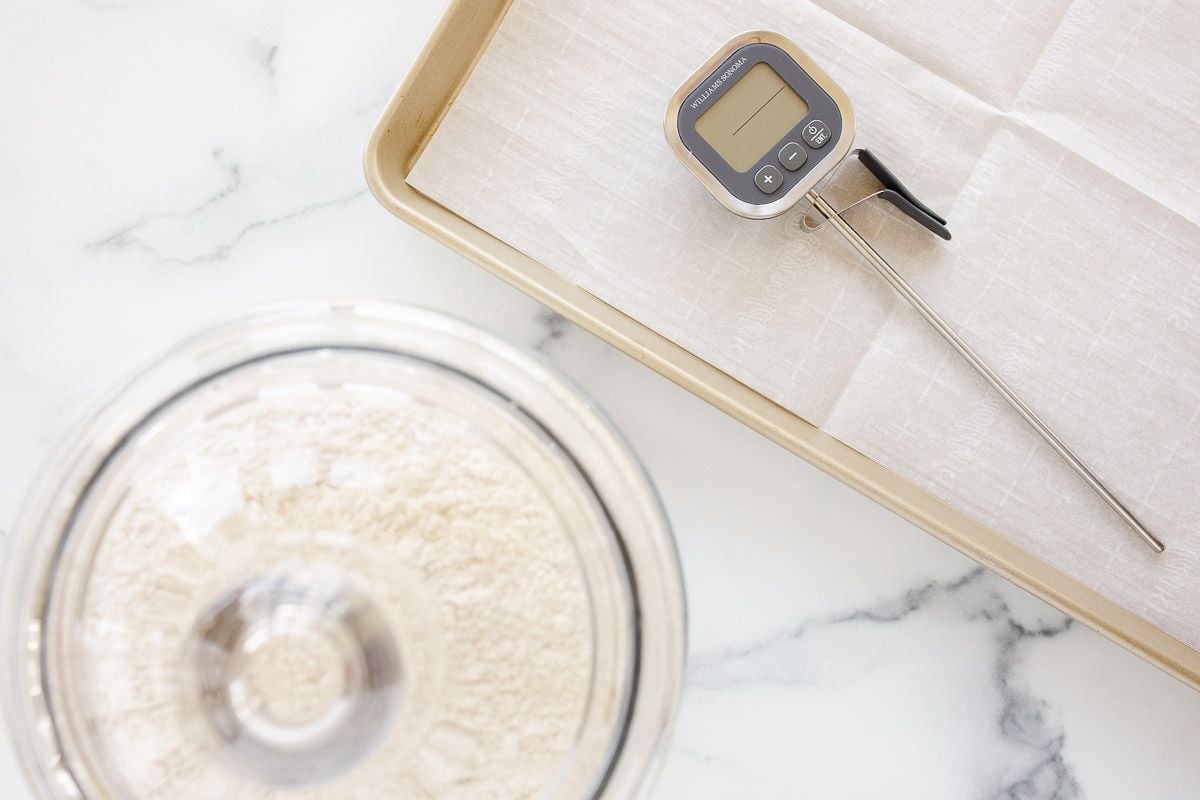 A glass canister full of flour, with a baking sheet and a digital thermometer to the side