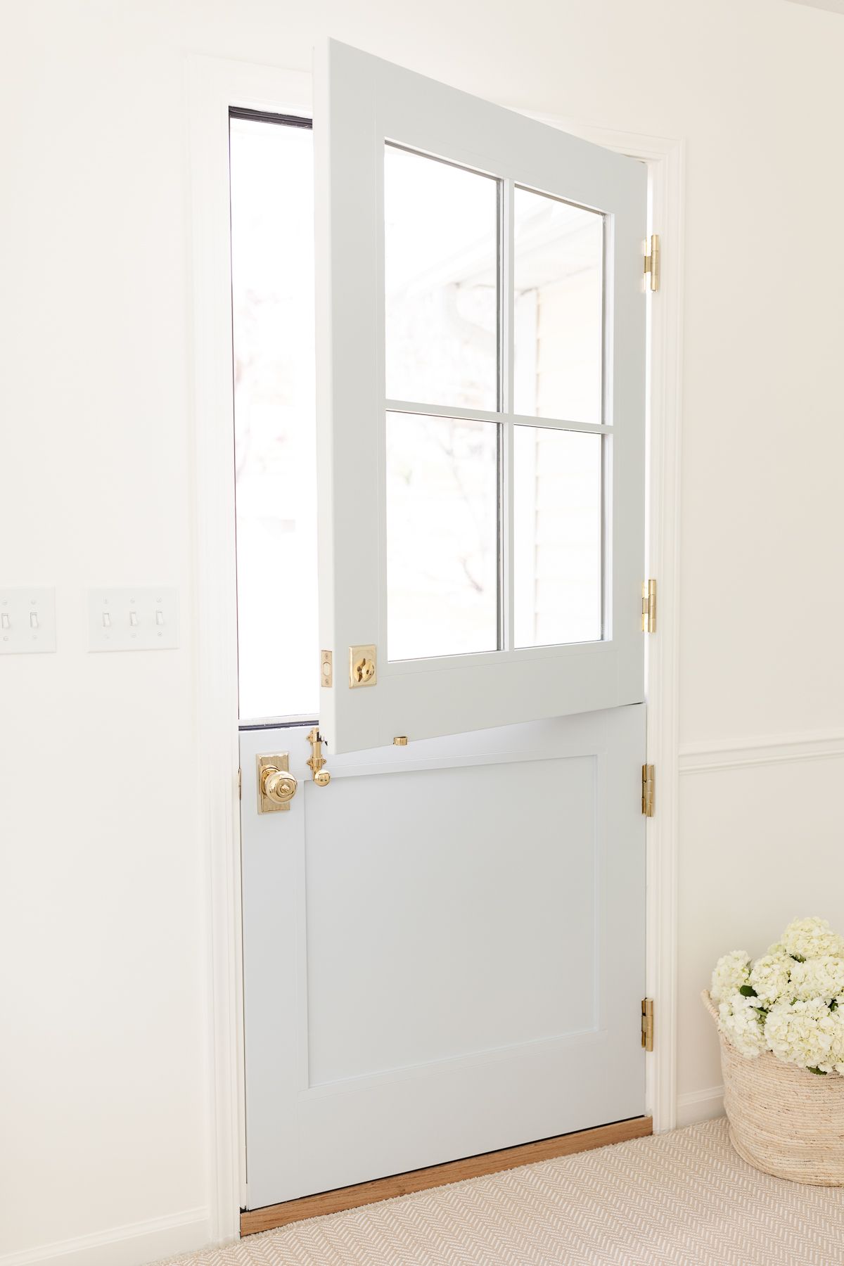 A soft blue Dutch door, top half open, with a basket of hydrangeas in an entryway, placed on top of a beige rug on carpet.