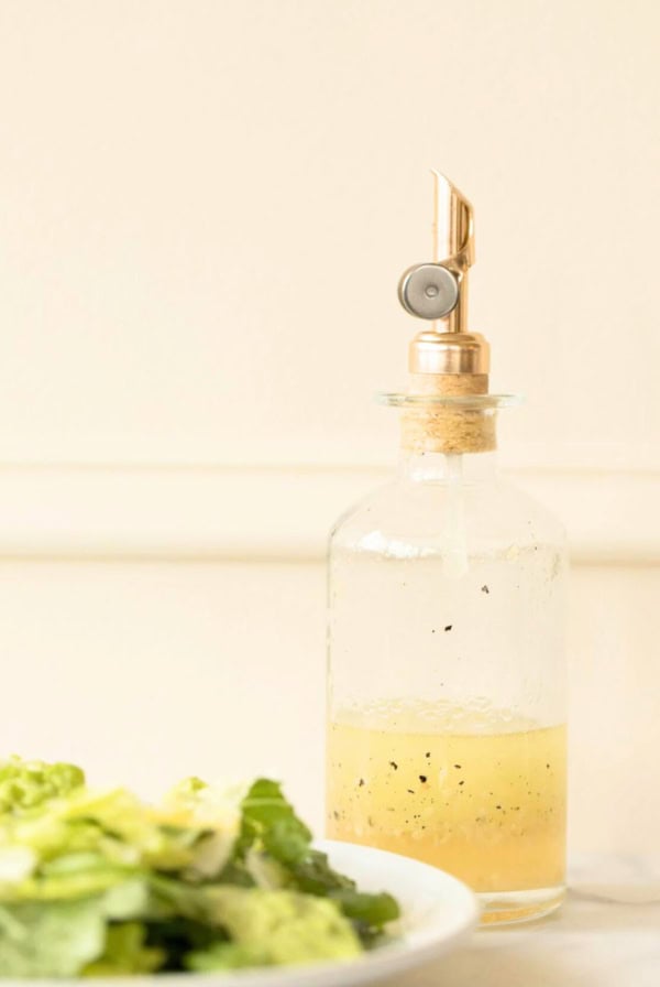 A glass bottle with a metallic spout contains a white wine vinaigrette. A bowl of fresh lettuce salad is seen in the foreground on the left side.